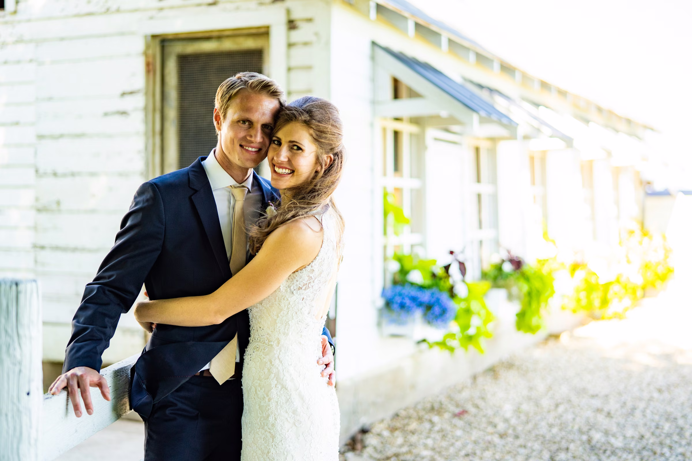 Katie embraces Tobias beside old white farm windows with soft foreground detail