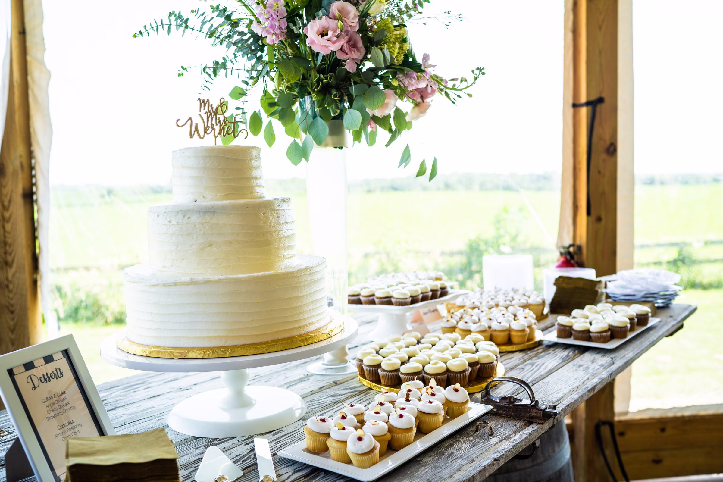 White wedding cake and rows of small desserts beneath a hanging floral arrangement