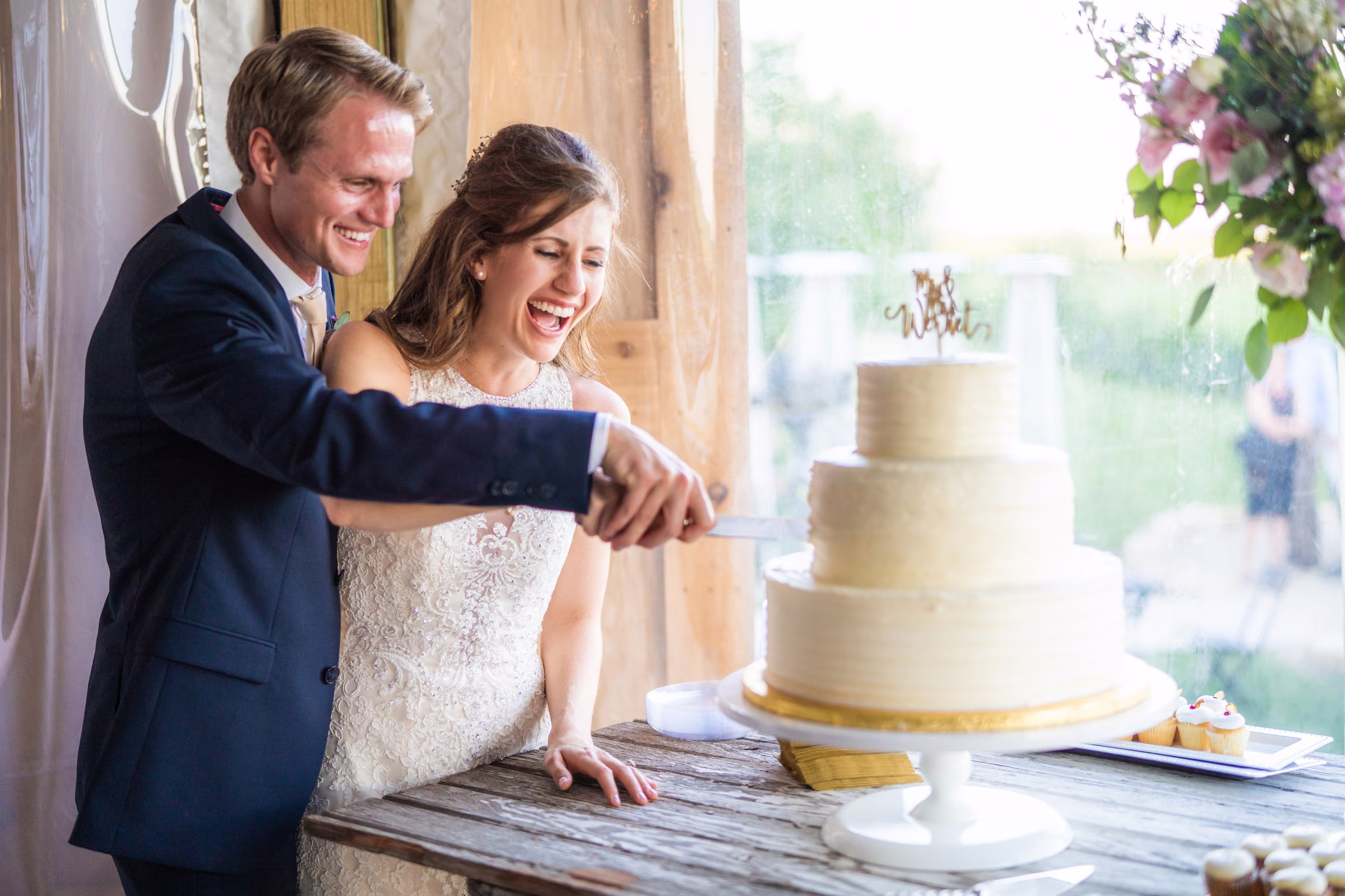 Katie and Tobias laugh together while cutting their tiered white wedding cake