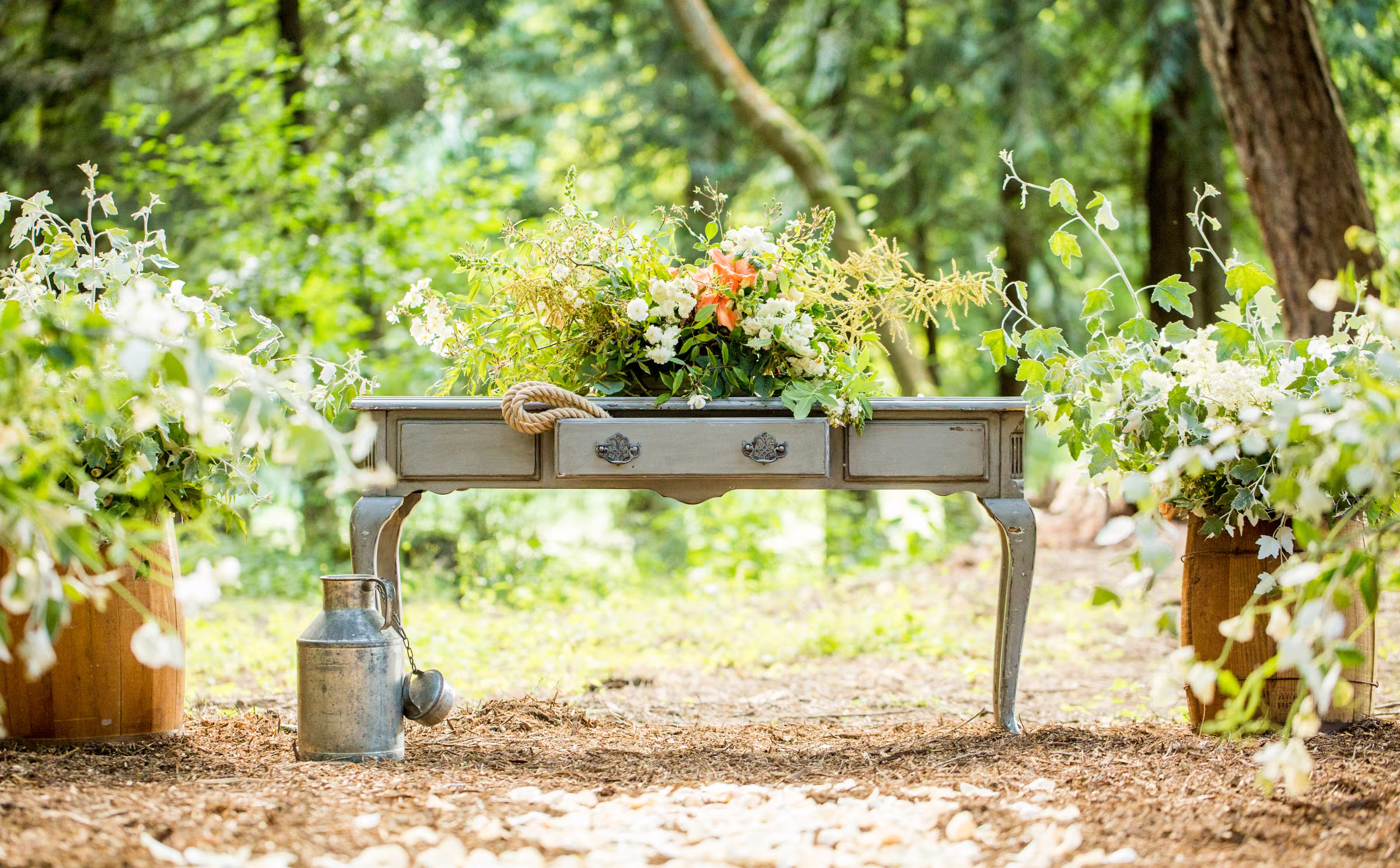 A small flower-covered table and simple ceremony details sit among trees beside the barn