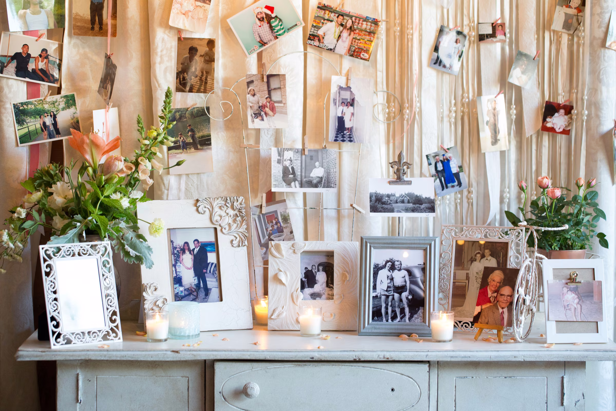 Old and recent family photographs hang above a white cabinet with flowers and candles