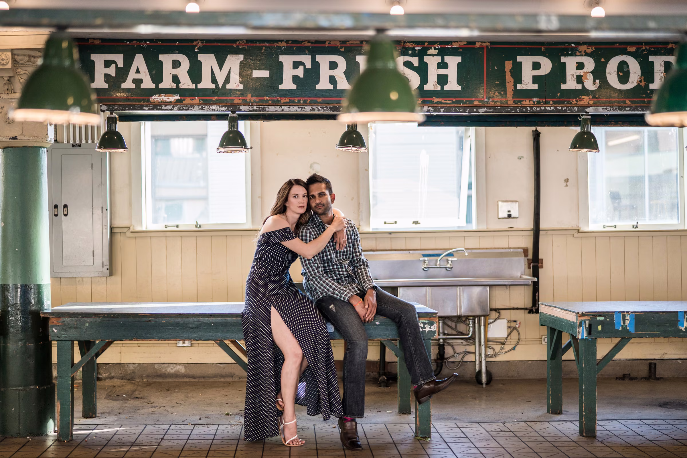 Kohleen and Kash sit close together on a wooden table beneath a farm-fresh produce sign at a Seattle market