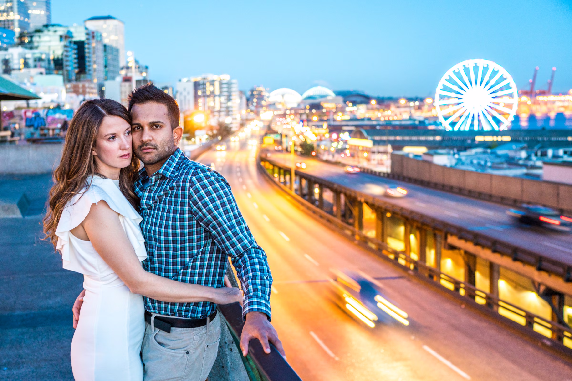 Kohleen and Kash stand close together above Seattle’s waterfront with the Ferris wheel and evening traffic behind them