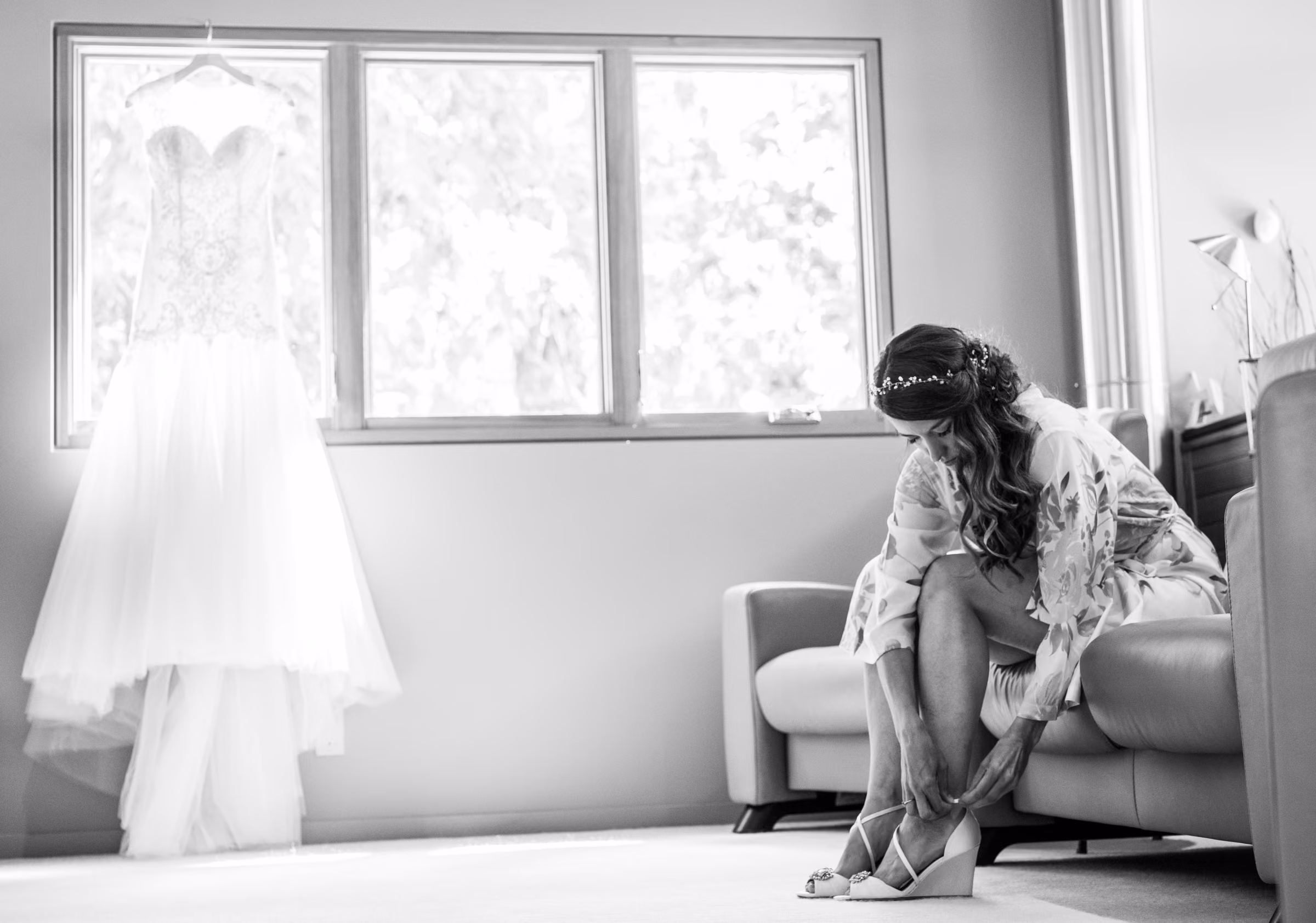 Kohleen puts on her wedding shoes by a window while her dress hangs nearby