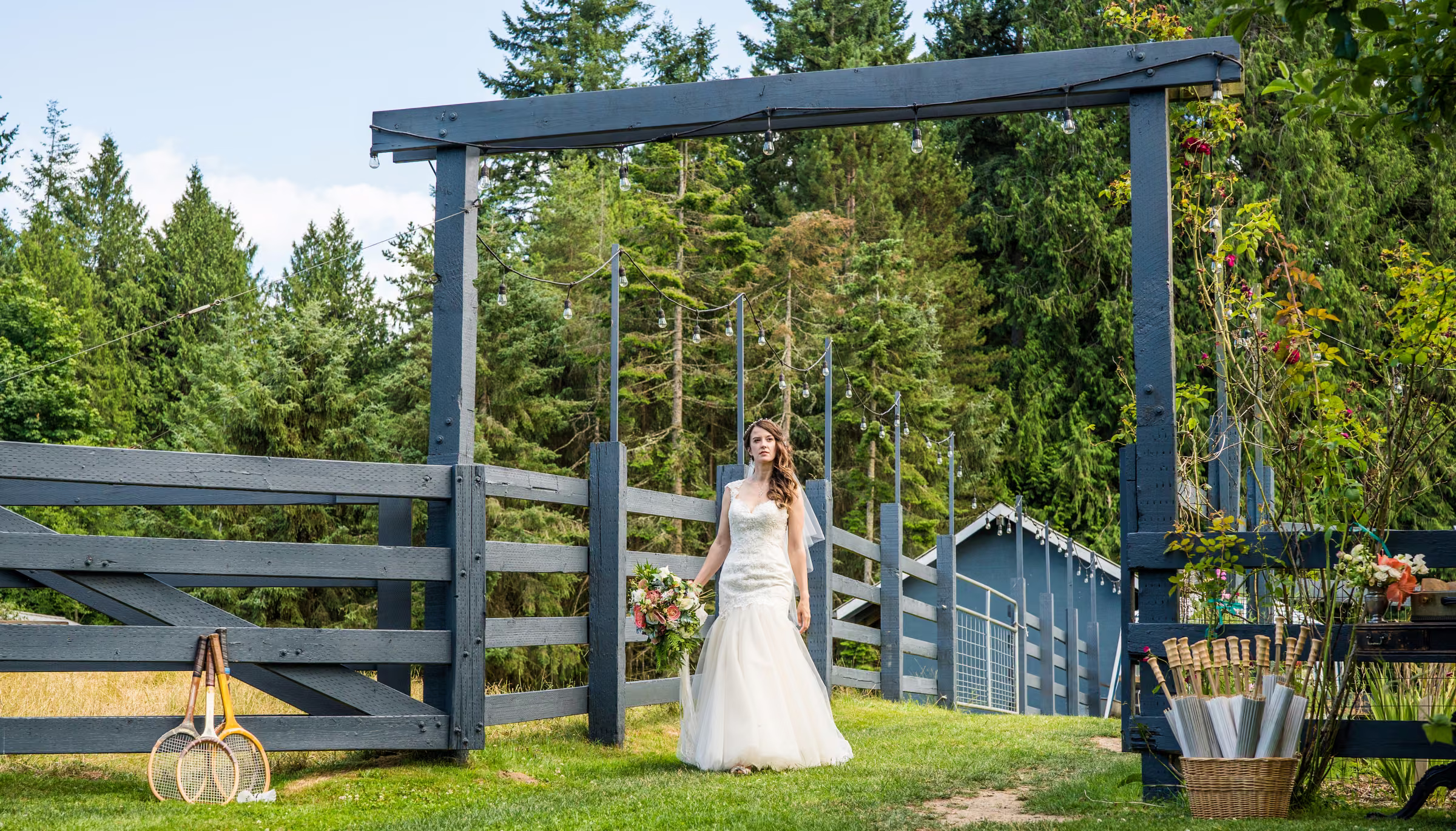 Kohleen stands in her wedding dress beside a blue gate with the farm and trees around her