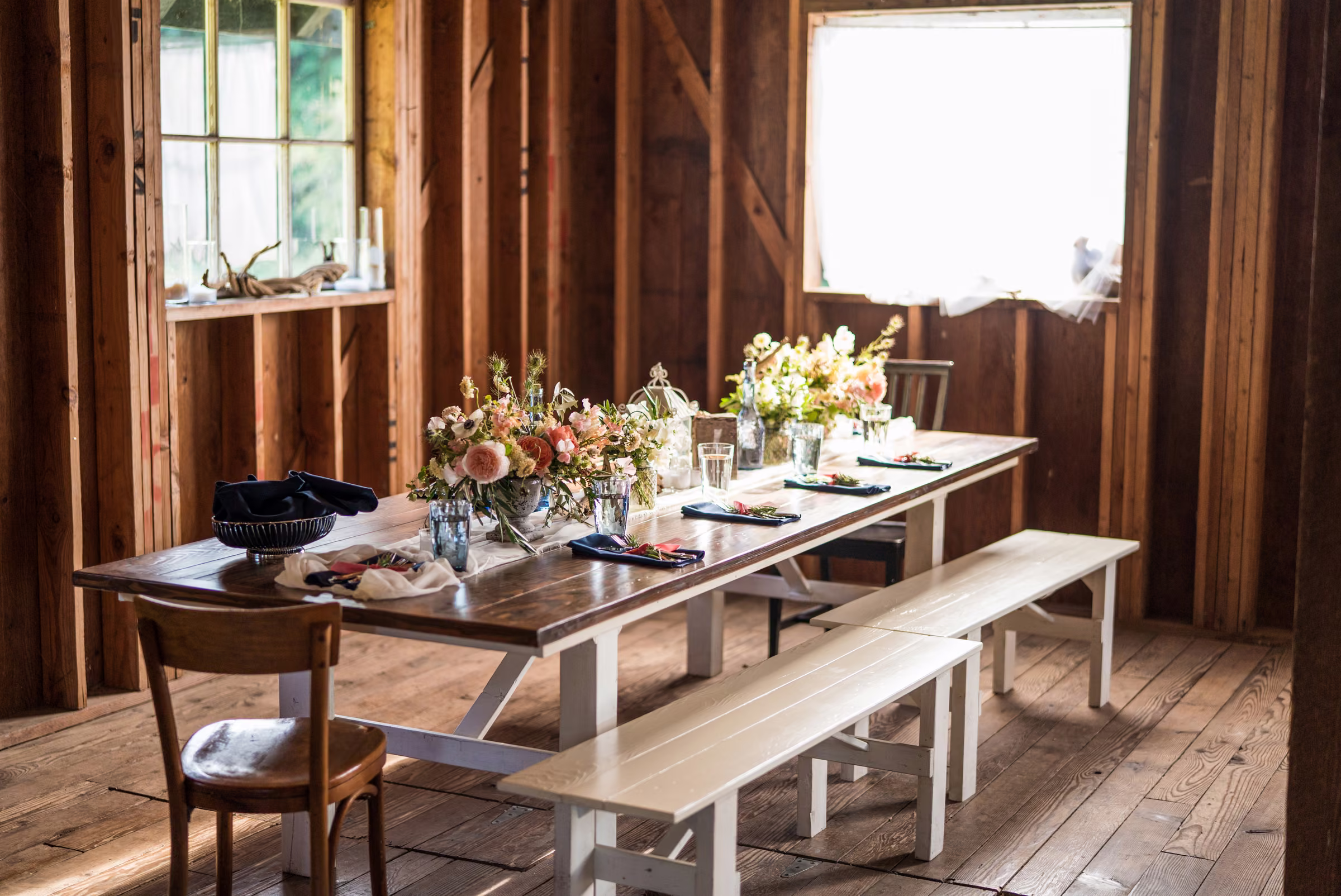 A long wooden reception table is set with flowers, place settings, and benches inside the barn
