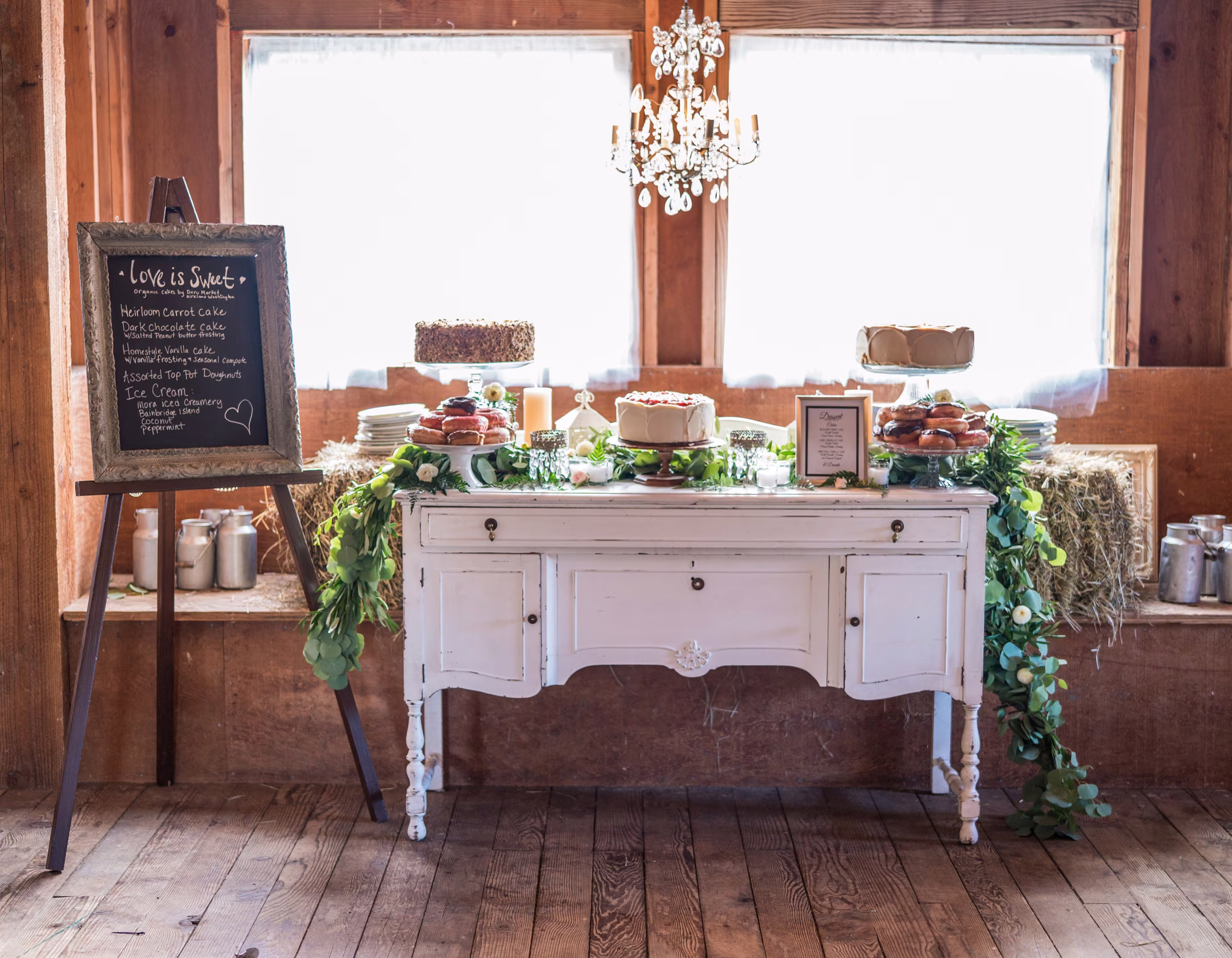 Wedding cakes and desserts are displayed on a vintage white table inside the barn