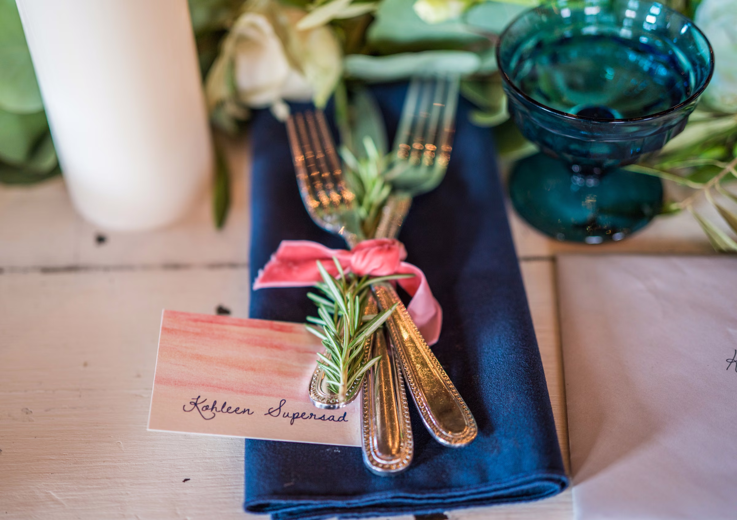 A place card reading Kohleen Supersad rests beside gold cutlery, a navy napkin, and blue glassware