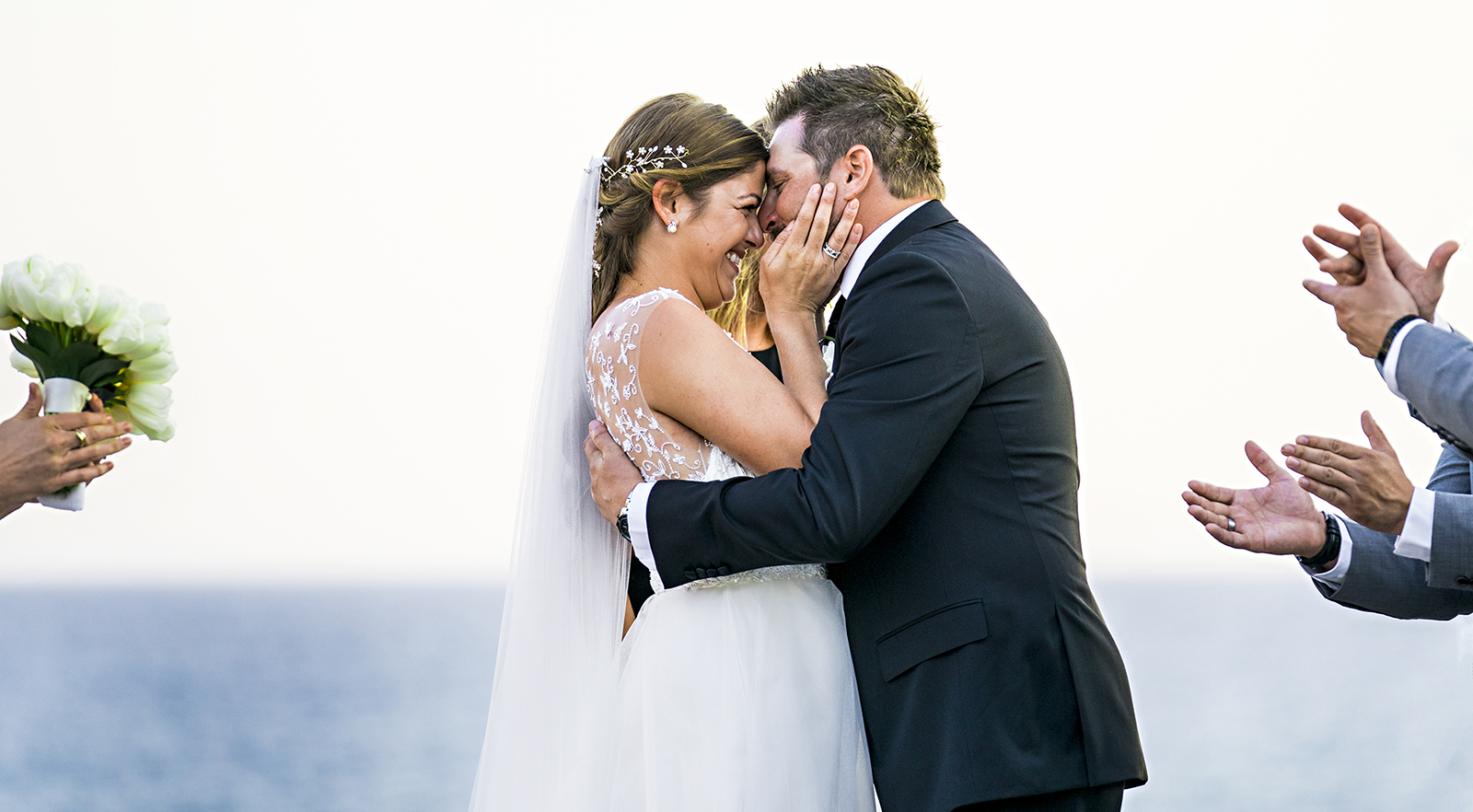 Newlyweds kiss with the hands of their wedding party close beside them
