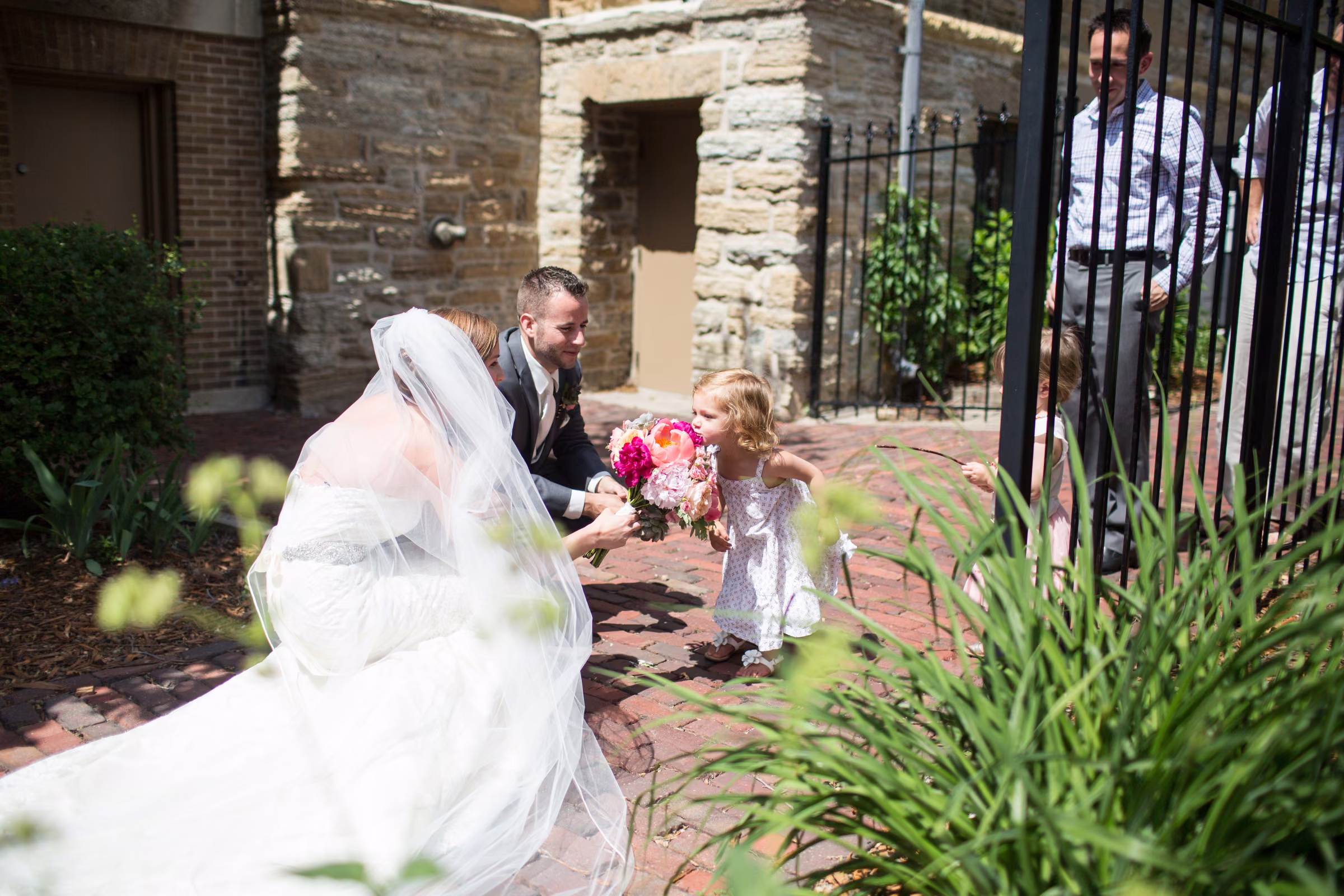 A flower girl leans toward Annie’s bouquet while Matt and Annie stop beside her on a brick path