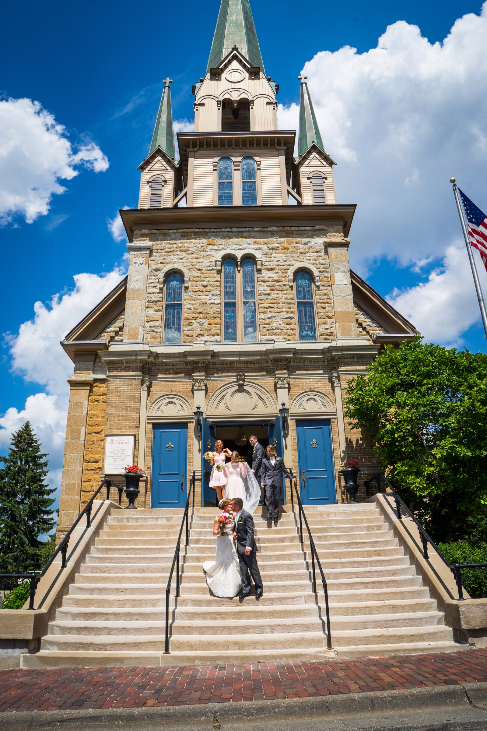 Matt and Annie kiss on the steps as they leave Our Lady of Lourdes Catholic Church beneath a blue sky
