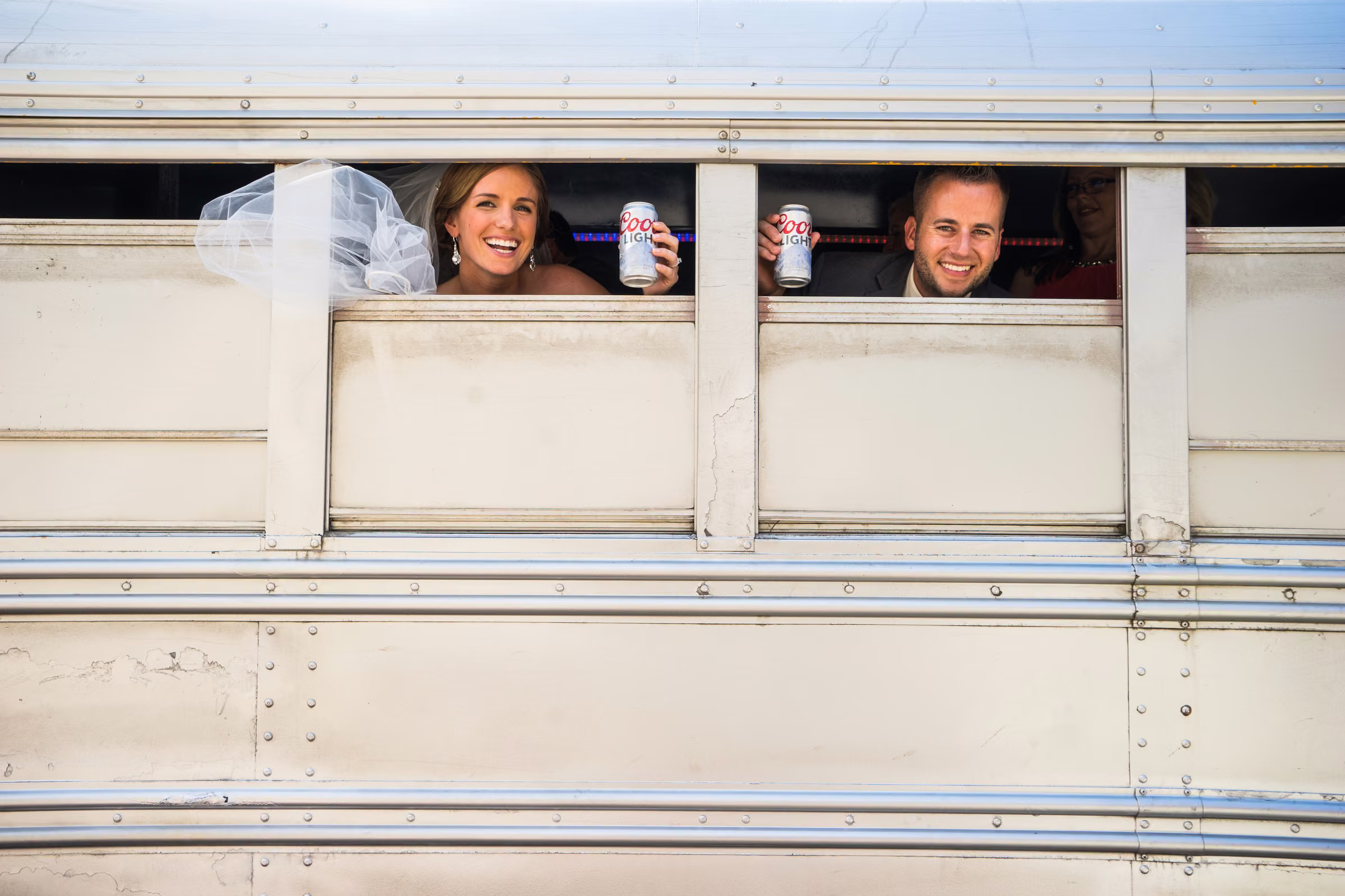 Annie and Matt smile out the windows of their wedding party bus holding drinks