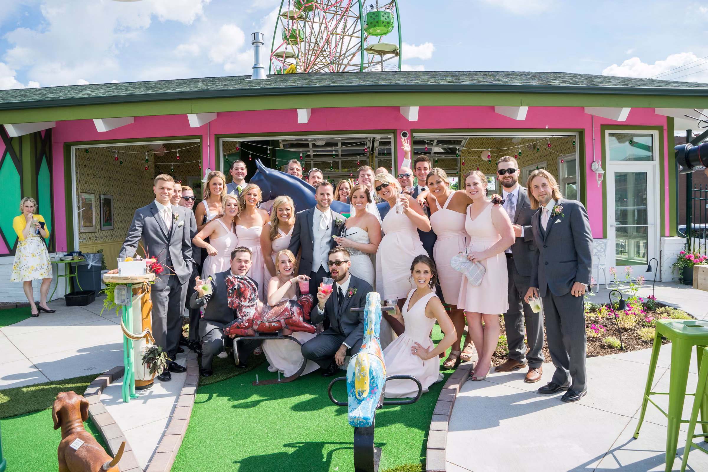 Matt, Annie, and their wedding party pose among mini-golf animals outside the pink and green Betty Danger’s Country Club with its Ferris wheel behind them