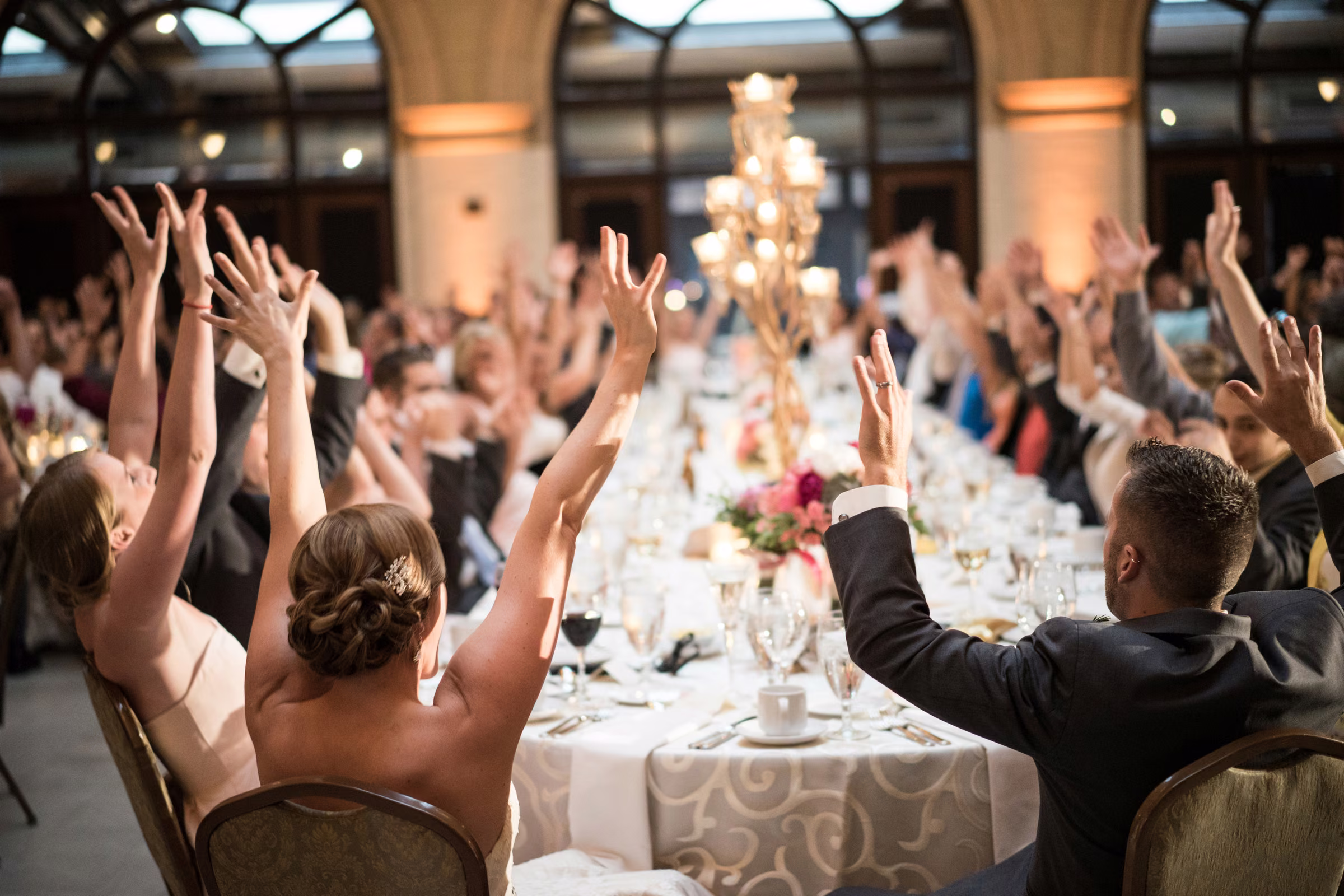 Wedding guests raise their hands around a long reception table at The Depot