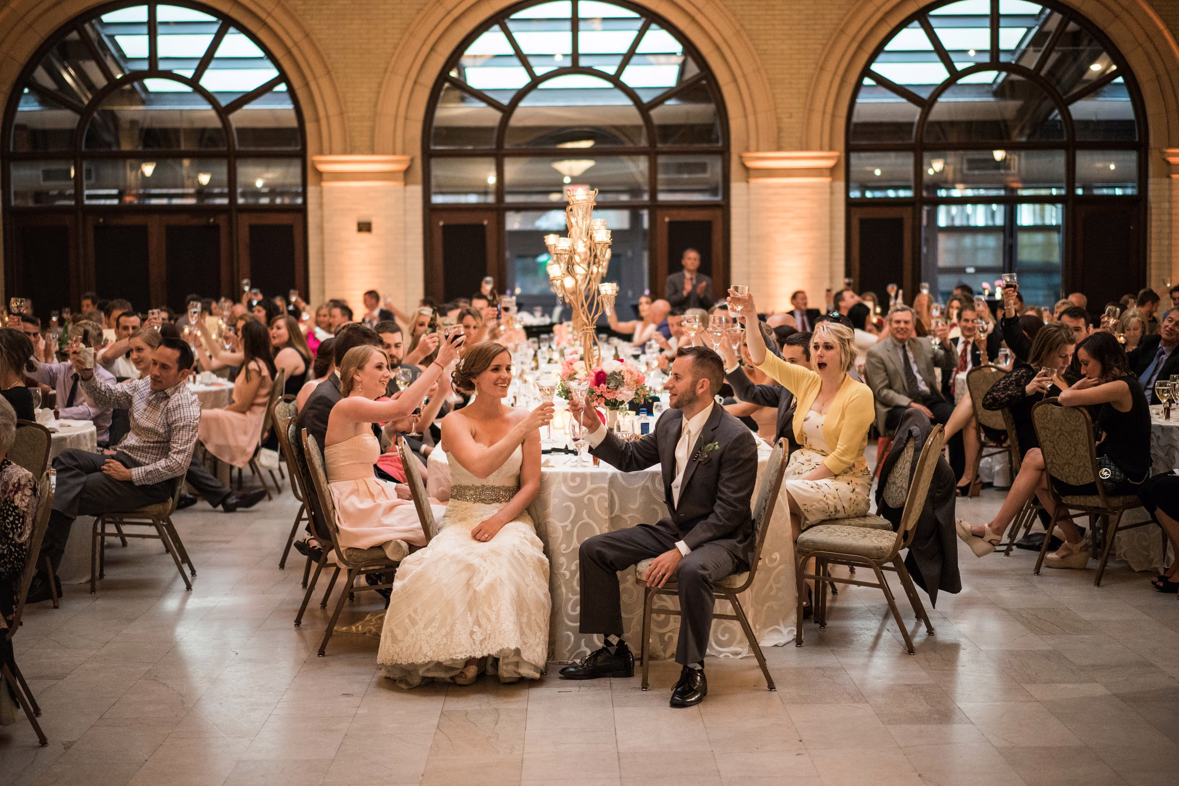 Matt and Annie sit among smiling guests raising glasses during a toast at The Depot